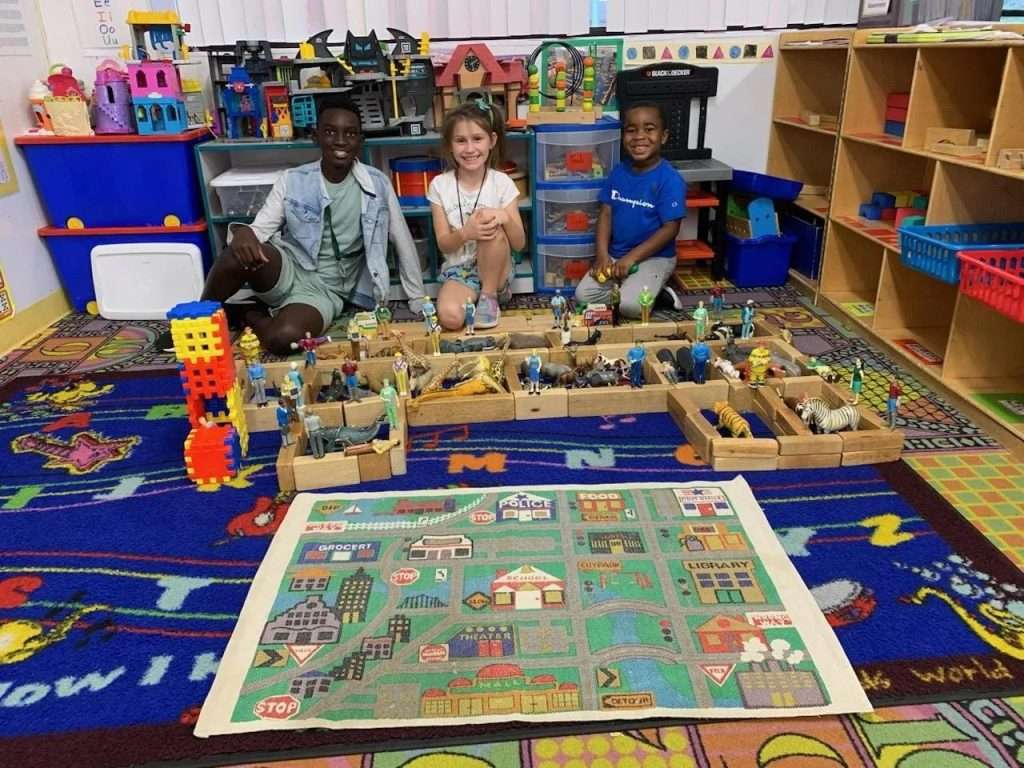 Three children sitting on a colorful rug surrounded by toy blocks and animal figures in a classroom.